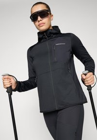 Black athletic jacket with zippered pockets, a fitted design, and a hood. Model holds trekking poles, wearing sleek black sunglasses.