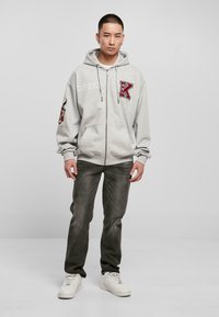 Haut zippé gris avec un grand logo « K », manches longues et cordons de serrage. Assorti à un jean gris foncé et des baskets blanches. Design simple.