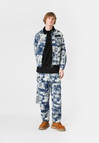 Chaqueta y pantalones de mezclilla de estampado azul y blanco, combinados con una blusa negra, que cuenta con bolsillos y una etiqueta. Calzado marrón completa el atuendo.
