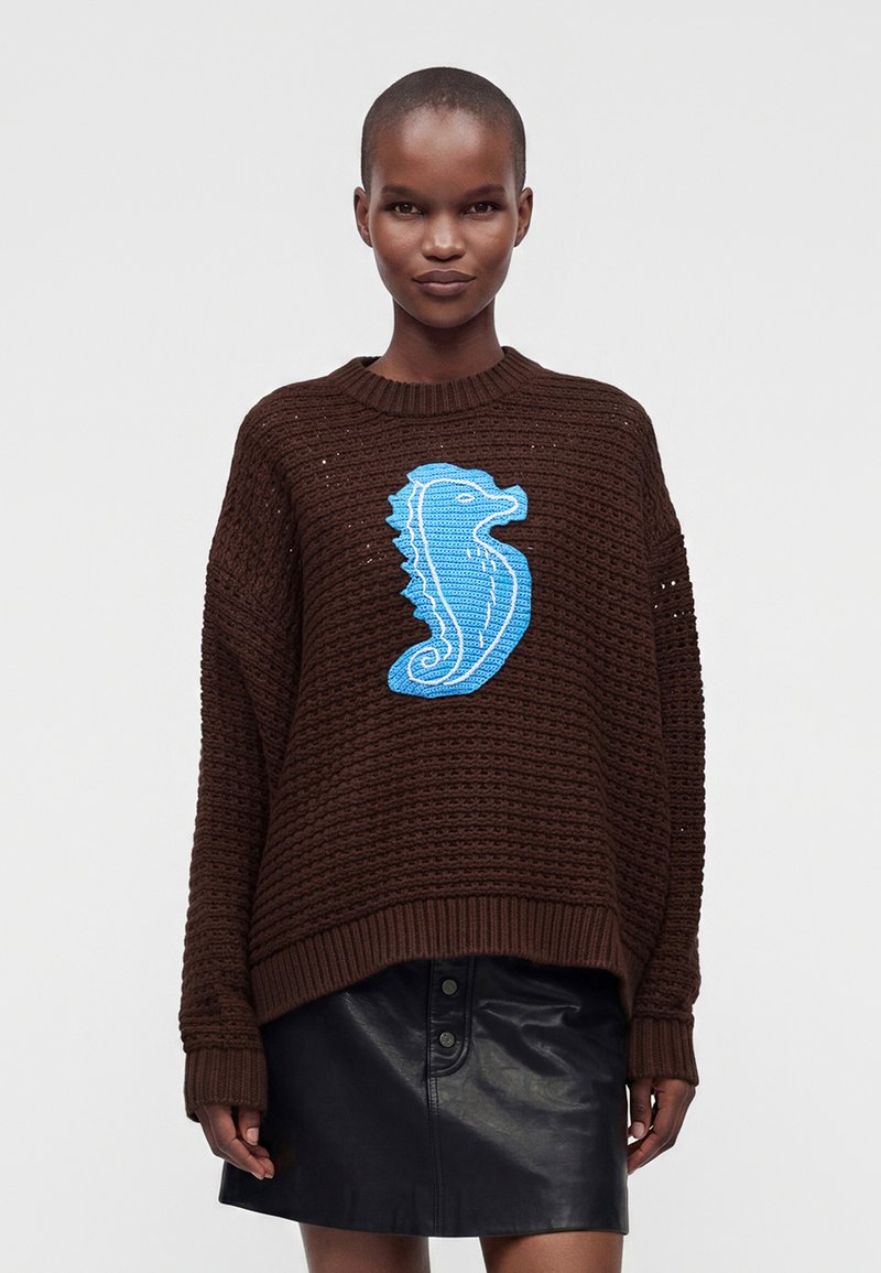 Personne portant un pull en laine marron avec un grand motif hippocampe bleu et une jupe en cuir noire sur un fond clair uni.