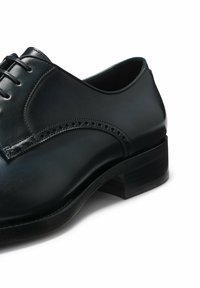 Scarpa da uomo in pelle nera con punta rotonda, dettaglio decorativo di brogue, chiusura con laccio e tacco basso in legno impilato. Texture liscia.