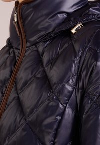 Mörkblå quiltad puffjacka med hög krage, brun dragkedjes trim och detaljer i guld. Slät yta med diamantformad sömnad.
