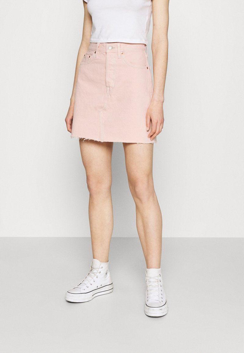 Femme portant une mini-jupe en jean rose clair avec un ourlet effiloché, un t-shirt blanc court et des baskets blanches montantes, debout sur un fond uni.