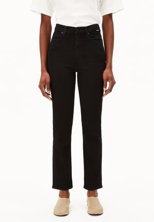 Person iført sorte bukser med høj talje og forlommer, kombineret med beige slip-on sko og en hvid kortærmet skjorte, stående oprejst.