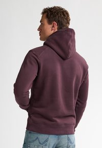 Burgunderfarbener Kapuzenpullover mit glatter Textur und gerippten Bündchen, kombiniert mit hellblauen Jeans. Verfügt über einen lockeren Schnitt und eine verstellbare Kapuze.