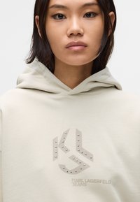 Hanorac bej deschis dintr-un material moale, având un logo texturat cu ace de argint. Include un glugă cu șnur și manșete îndoite.