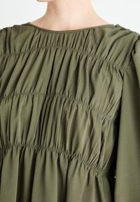 Robe rassemblée vert olive présentant un tissu léger et lisse, avec un col rond et des détails subtils élastiqués le long du corsage.