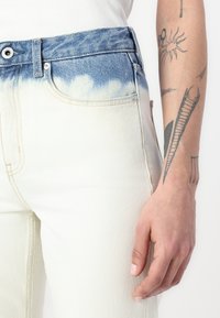 Calças de ganga de cor creme clara com efeito de tingimento azul na cintura, apresentando um corte reto e design clássico de cinco bolsos.