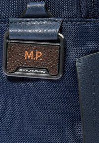 Détail d'un sac en nylon texturé bleu marine avec une étiquette en cuir marron foncé embossée des initiales "M.P." et des accents métalliques avec la marque.