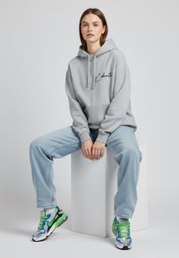 Felpa grigia con stampa nera "Liberté", abbinata a jeans baggy azzurri e sneakers Nike multicolori con dettagli verdi.