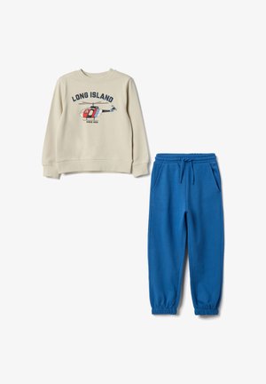 Sweat-shirt beige avec graphique "LONG ISLAND" et motif d’hélicoptère ; associé à un pantalon de jogging bleu à taille élastique avec chevilles resserrées.