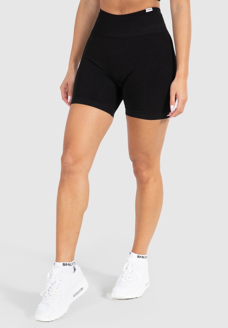 Kobieta ubrana w czarne szorty rowerowe z wysokim stanem, białe sneakersy i pasujące białe skarpetki kostkowe na jednolitym tle.