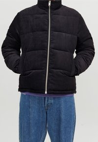 Czarna kurtka puchowa z sztruksu z wysokim kołnierzem i srebrnym zamkiem błyskawicznym, posiadająca poziome pikowane sekcje i elastyczne mankiety, noszona na fioletowej bluzce.