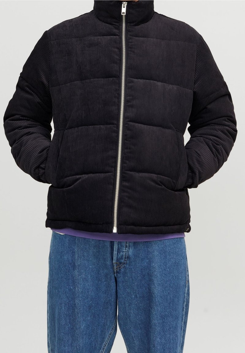 Czarna kurtka puchowa z sztruksu z wysokim kołnierzem i srebrnym zamkiem błyskawicznym, posiadająca poziome pikowane sekcje i elastyczne mankiety, noszona na fioletowej bluzce.