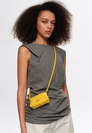 Kobieta z kręconymi włosami, ubrana w szary bezrękawnik i kremowe spodnie, nosząca małą żółtą torebkę na pasku przewieszoną przez ramię z długim paskiem.