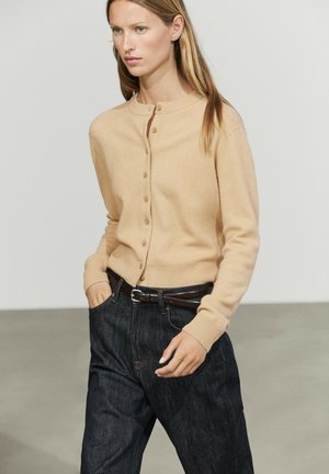 Donna che indossa un cardigan beige con bottoni e jeans scuri con cintura nera, in piedi con una mano in tasca su uno sfondo neutro.