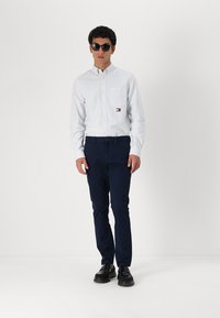 Wit gestreepte overhemd met knopen en een klein logo, gecombineerd met marineblauwe broek. Draagt zwart zonnebrillen en zwarte schoenen.