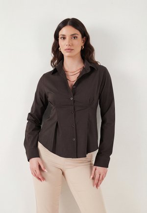 Kvinde med langt bølget mørkt hår bærer sort knapbluse, lagdelte guldhalskæder, gyldne øreringe og beige bukser, står op ad en hvid væg.