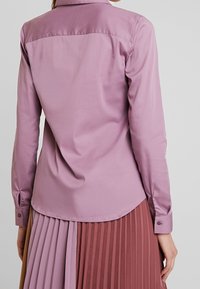 Donna che indossa una camicia a maniche lunghe di lavanda e una gonna plissettata con pannelli verticali in marrone, lavanda e rosa scuro.