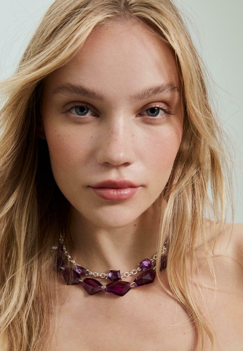 Lilla gemstone halskæde med forskellige former og størrelser, prydet med en sølvkæde. Glatte teksturer og skinnende overflader skaber visuel interesse.