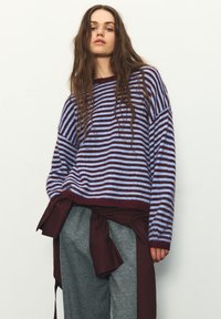 Pruhovaný pletený sveter v svetlomodrej a bordovej farbe, s voľným strihom, širokými rukávmi a rebrovaným lemom; doplnený sivými teplákmi.