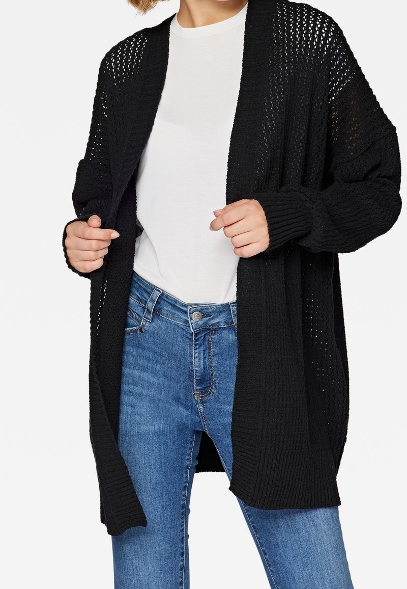 Cardigan noir en maille ouverte avec manches longues et poignets côtelés, porté par-dessus une chemise blanche et associé à un jean bleu. Tissu texturé et coupe ample.