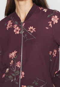 Burgundská bomber bunda s květinovou výšivkou v růžové a zelené. Má přední zip a žebrovaný límec, který vystavuje texturovanou látku.
