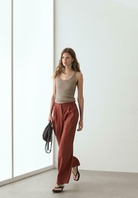 Femme marchant à l'intérieur portant un débardeur beige, un pantalon large couleur rouille, des sandales noires, et tenant un sac noir dans sa main gauche.