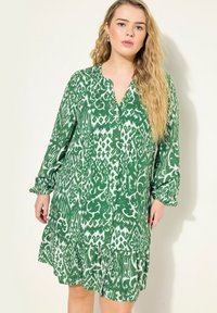 Robe verte à motif en tissu léger, avec un décolleté en V, des manches longues avec des poignets froncés, et un ourlet à volants.
