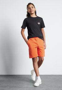 Svart bomullströja med en liten grafik på bröstet, tillsammans med ljusorange shorts, båda med avslappnad passform. Vita sneakers och strumpor avslutar looken.