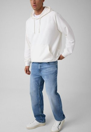 Hombre con sudadera blanca con capucha, vaqueros azul claro y zapatillas blancas, de pie contra un fondo liso gris claro.