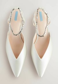 Valkoinen satiininen slingback-kengät, joissa on kärjellinen muoto ja hopeaketju yksityiskohtana nilkan ympärillä. Sileä rakenne ja solakka siluetti.