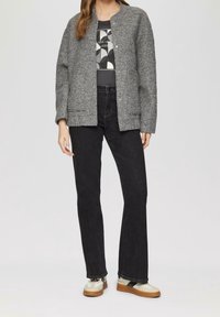 Giacca grigia con texture, maniche lunghe, chiusura a bottone, abbinata a pantaloni neri a zampa d'elefante e scarpe da ginnastica con una combinazione di colori crema e marrone.