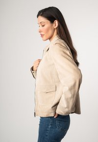 Beige Wildlederjacke mit Reißverschluss, seitlichen Taschen und Kragen. Strukturierte Oberfläche, taillierte Passform und minimalistische Nähdetails.