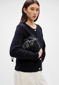 Juodas kardiganas su tekstūru, apvaliu iškirpimu ir perlų sagomis, derinamas su juoda plisuota rankine. Modelis stovi prieš šviesią fono.