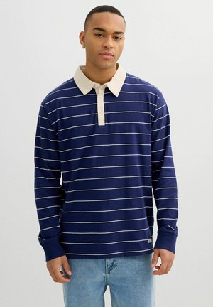 Hombre con polo de manga larga azul marino con rayas horizontales blancas y cuello, combinado con vaqueros azul claro, sobre fondo liso.