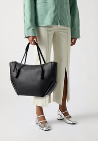 Sac fourre-tout en cuir noir à forme arrondie, avec deux poignées supérieures et une fermeture éclair ; doté d'une texture lisse et d'accents minimalistes.