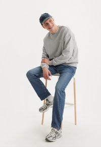 Szary sweatshirt z okrągłym dekoltem, niebieskie dżinsy i szare buty sportowe. Osoba siedząca na drewnianym stołku, uśmiechająca się, nosząca niebieską czapkę.
