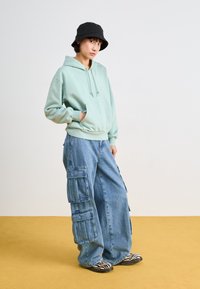 Hoodie vert menthe avec une poche avant, associé à un pantalon cargo large bleu clair avec plusieurs poches. Chapeau seau noir et chaussures rayées.