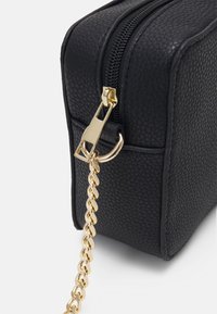 Angolo di una borsa nera in pelle strutturata con zip dorata e catena spessa dorata attaccata su sfondo bianco.