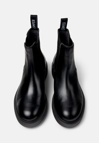 Une paire de bottines Chelsea en cuir noir avec des semelles épaisses et des languettes de tirage, étiquetées "Camper", présentée en vue de dessus sur fond blanc.