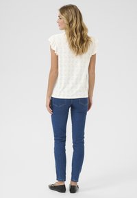 Haut blanc à détail en œillet avec de courtes manches volantes, associé à un jean skinny bleu et des chaussures plates noires, vu de dos.