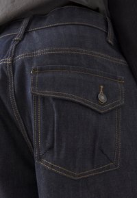 Dark blue denim jeans featuring a back pocket with a distinctive metal button and contrasting yellow stitching along the seams.