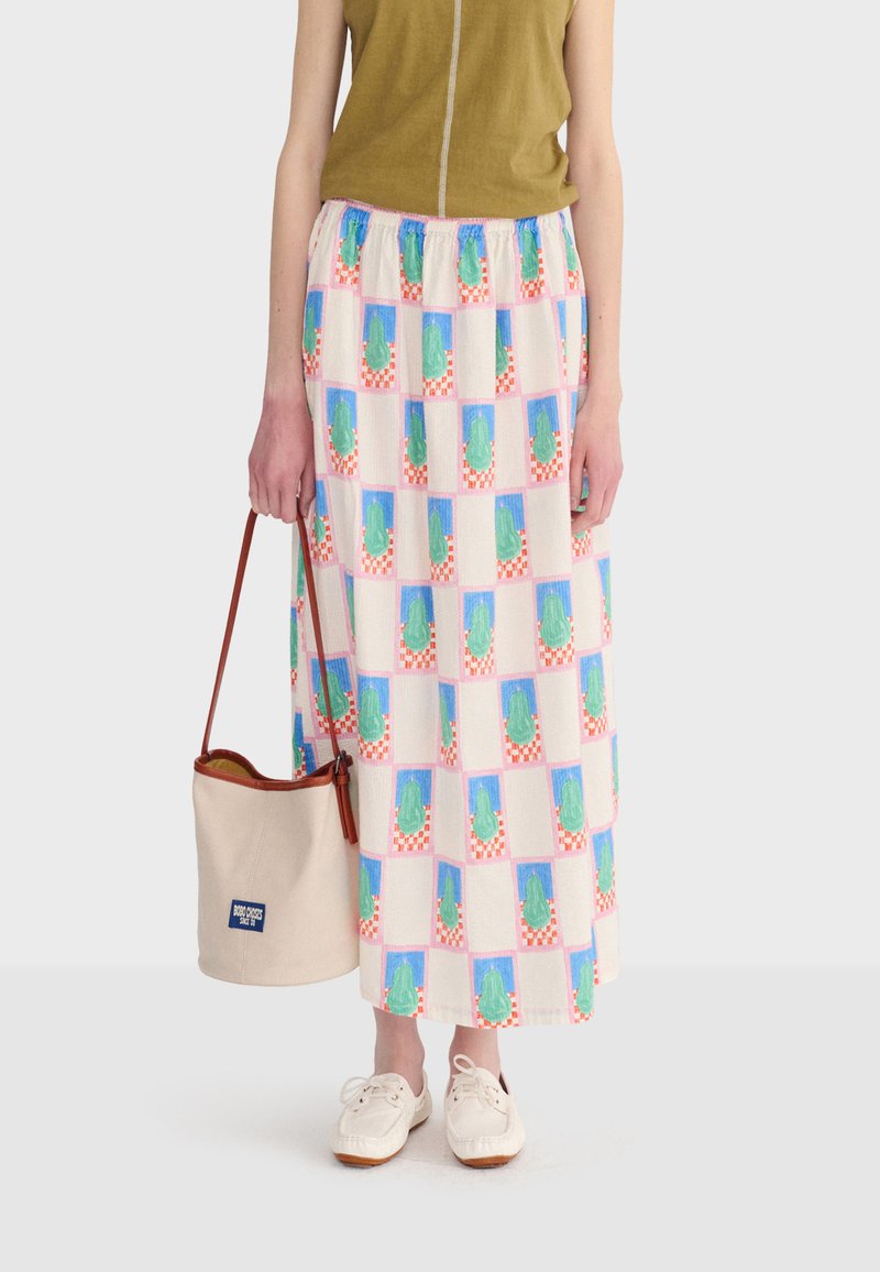 Personne portant un haut vert sans manches, une jupe longue à motifs avec des imprimés de cactus verts, des chaussures blanches, tenant un sac seau beige avec des anses marron.