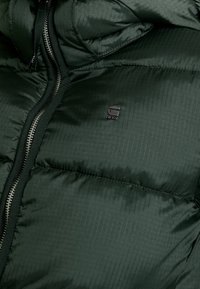 Mörkgrön quiltad pufferjacka med en synlig silverfärgad dragkedja och en liten "RAW"-logotyp på vänster bröstområde.