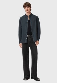 Camicia scura con colletto a punta, indossata sopra una maglietta nera; abbinata a jeans neri a gamba larga e calzature nere. Design semplice e pulito.