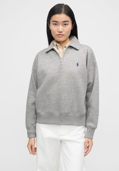 Γυναίκα που φοράει γκρι φούτερ με φερμουάρ μέχρι τη μέση πάνω από μπεζ ριγέ πουκάμισο και λευκό παντελόνι, στέκεται μπροστά σε απλό φωτεινό φόντο.