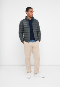 Grå pufferjakke med vandret syninger, lynlås foran, parret med en navy sweater og lyseblå skjorte, beige bukser og hvide sneakers.