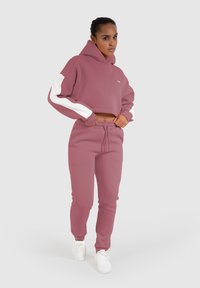 Rosa bauchfreie Kapuzenjacke mit weißen Ärmeln und passenden Jogginghosen. Hergestellt aus weichem Stoff, bietet eine legere Passform und eine Kordelzug-Taille.