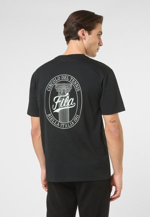 Mann trägt schwarzes T-Shirt mit weißem, rundem Fila-Logo und Säulengrafik auf der Rückseite, steht vor einfarbigem grauem Hintergrund.
